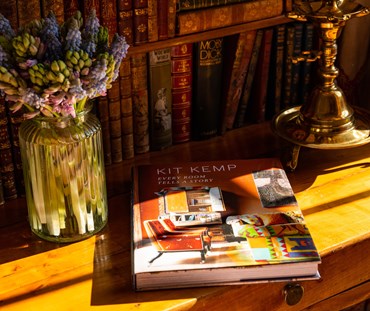 Kit Kemp's Every Room Tells a Story book, shown on antique desk, next to a vase of flowers, and a brass lamp base.