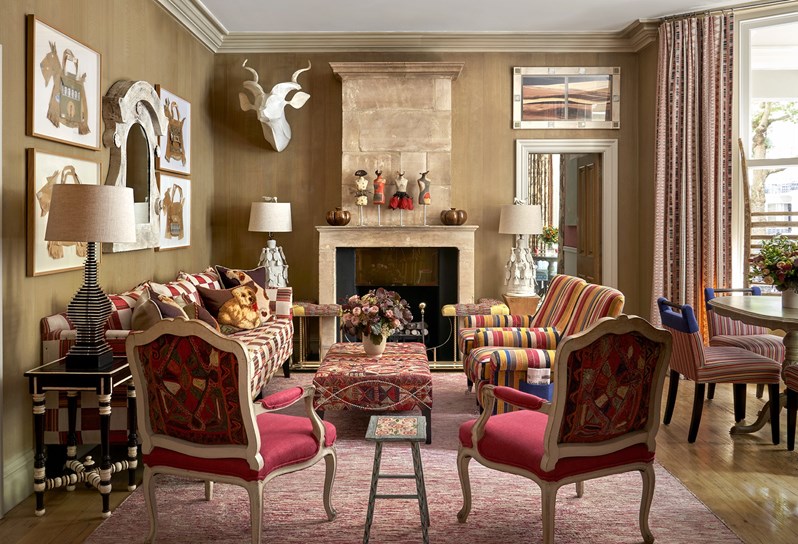 Wide shot of the Drawing Room at Knightsbridge Hotel, the room displays a sofa on the left, in the foreground the backs of two arm chairs that face into the room. in the centre a low embroidered coffee table, on the right hand side of the seating area, two striped arm chairs. On the fart right of the image we see a round dining table with 4 striped chairs around it.