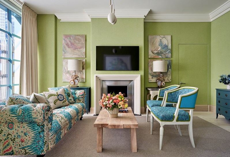 A green walled sitting room. Two blue and green Whitby chairs on one side of a coffee table, and a blue and green floral patterned sofa on the opposite side. A fireplace and TV are on the centre of the wall, and two square prints are hung above a table and lamp either side.