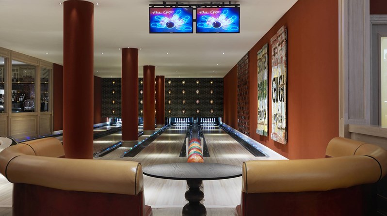Lounge area consisting of half moon shaped leather sofas in bright colors and a wooden side table in front of two bowling lanes with bright colored bowling balls and intricate artwork on the walls.