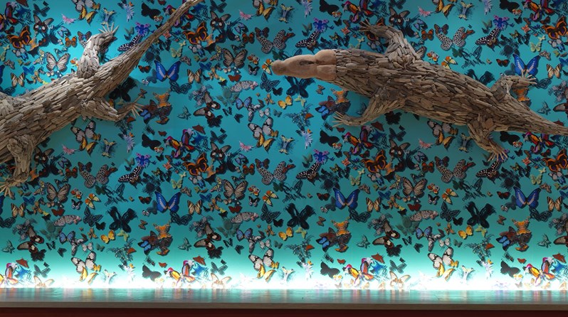A bright turquoise wall with butterflies in various colors, topped with two large wooden sculptures of crocodiles crawling across the wall.