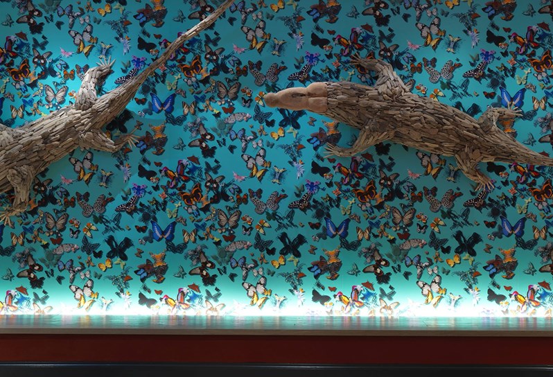 A bright turquoise wall with butterflies in various colors, topped with two large wooden sculptures of crocodiles crawling across the wall.