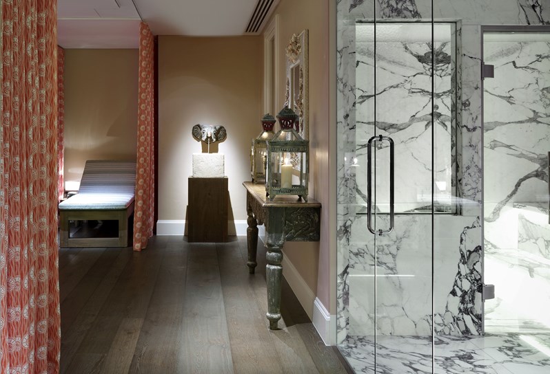 Look through onto the spa relaxation area. There are orange and white curtains hanging from the ceiling and a white relaxation bed. On the right you can see the marble steam room. At the very end of the room there is a small table with an elephant shaped fountain.