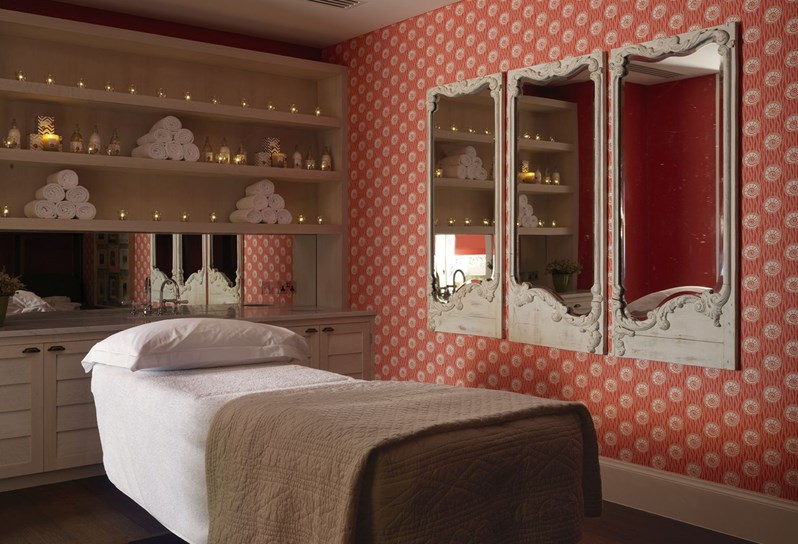 Spa treatment room with massage bed, on the bed is a white fluffy pillow and a blanket. On the right wall there is orange and white wallpaper. There are three framed mirrors hanging on the wall. On the back wall in the middle there are shelves with fresh white towels and Rik Rak products.