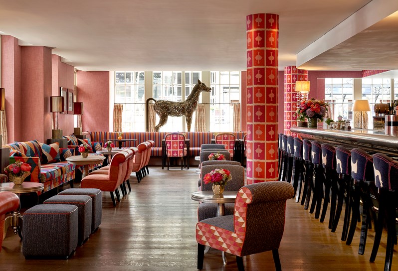 A wide shot of the bar area at Brumus in Haymarket Hotel. On the right, there is a selection of low bar tables and seating. On the left, a row of bar stools are lined up in front of the bar.
