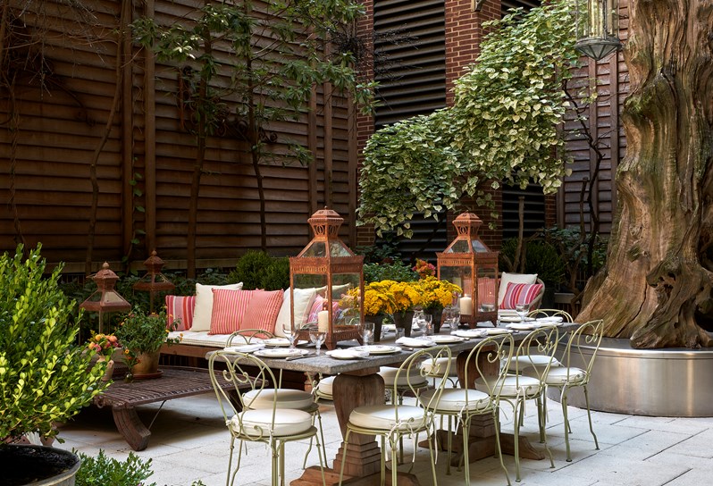 Shot from the curtain wall of the bar, of a large stone table set up for ten guests in the sculpture garden at the Crosby Street Hotel with lounge sofas at the back of the shot. There is a large tree on the back right