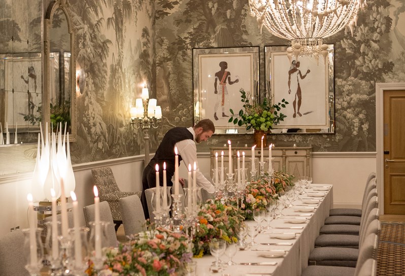 A portrait shot looking down a long table dining set up in the Shooting Gallery at Haymarket Hotel. The table is dressed with colourful bouquets of flowers and long candle sticks. At the far end a waiter is laying a place setting.