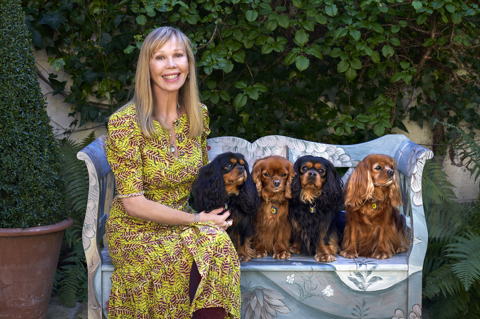 An mage of kit kemp sitting on a painted blue bench in a yellow patterned dress with her four King Charles Caviller spaniels. 