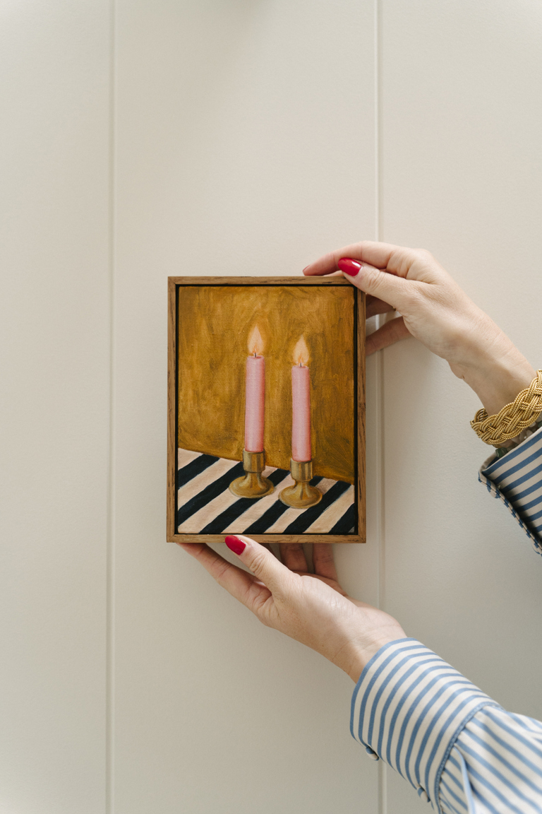 Two hands of a woman placing a small framed artwork by Domenica Marland on a white wall.