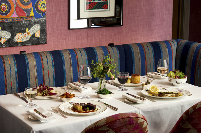 A table of four laid with main dishes accompanied by glasses of wine at Brumus. The table is positioned in front of banquette seating with art in partial view hanging on the wall