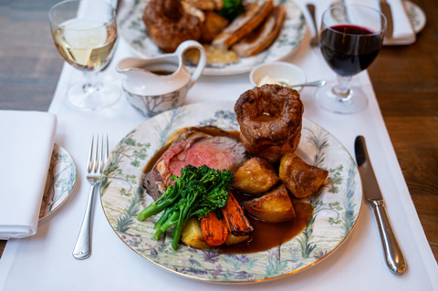 Sunday Roast at The Potting Shed. A plate of roast beef, with Yorkshire pudding, potatoes, vegetables and gravy with a plate of roast pork out of focus opposite. Both roasts are paired with glasses of wine