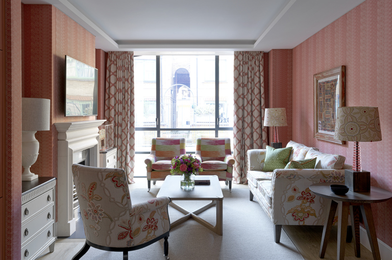 A wide shot of the living space are upholstered in a soft red fabric and a sofa, two armchairs and a coffee table frame a fireplace on the left hand side of the room. At the end of the room are floor-to-ceiling windows of the 9-11 Richmond Buildings.