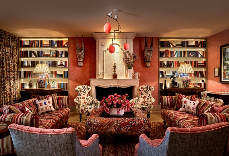 The Drawing Room at Warren Street Hotel with two sofas upholstered in a dark red striped fabric, a large rectangular footstool in a red embroidered fabric and two curvaceous statement armchairs in a cream, red and blue floral fabric. In the background is a stone fireplace with bookshelves on either side