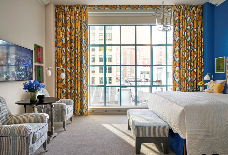 A room with floor to ceiling windows at the back that have yellow and blue curtains with a tropical fruit print. To the left, two blue and white striped armchairs and a coffee table underneath a TV and two green and pink prints on the wall. To the right, a bed is partially in view with a yellow and blue cushion and a blue and white ottoman at the end.