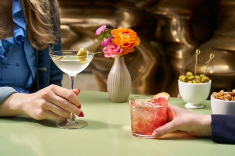 A wide shot of two people enjoying cocktails outside at Ham Yard. There are bar snacks of olives and nuts on the table.