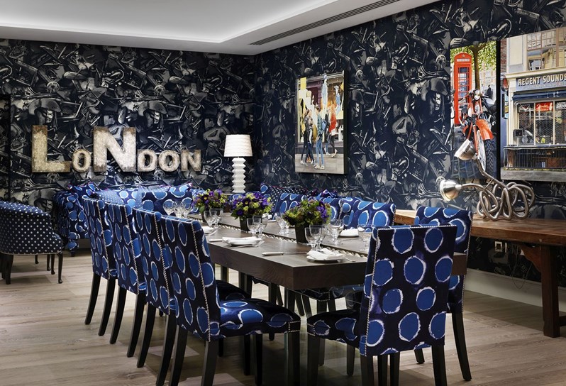 A table set for private dining for 10 people on comfortable blue patterned chairs, in the Archer private event space, featuring trendy transport inspired wallpaper by John Paul Gaultier in dark colors and quirky design pieces and artwork.