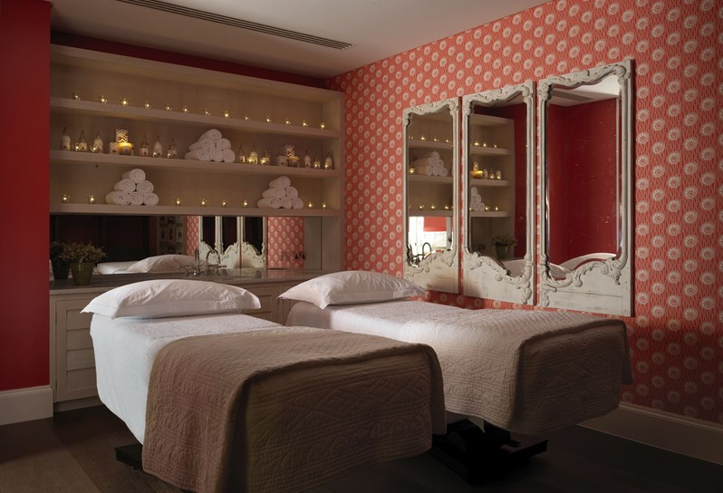 Double Spa treatment room with two massage beds, on each bed there is a white fluffy pillow and a blanket. On the right wall there is orange and white wallpaper. There are three framed mirrors hanging on the wall. On the back wall in the middle there are shelves with fresh white towels and Rik Rak products.