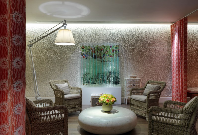 Spa relaxation area with four chairs with cushions in and a round stone table in the middle. There is a bouquet of flowers on the table. On the back wall there is a coulorful piece of art work. On the right there is a orange and white curtain hanging  from the ceiling. On the left is a large floor lamp.