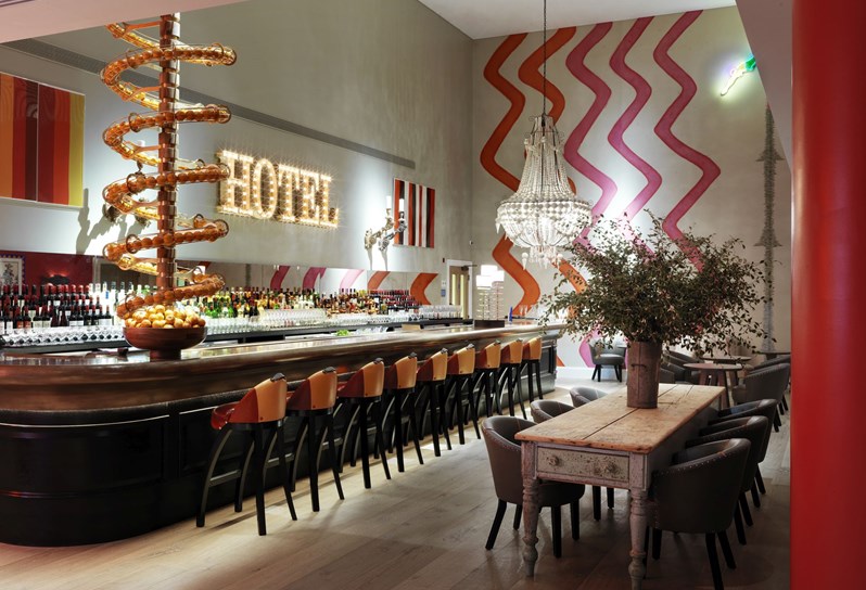 Angled view of a fully stocked bar with high chairs in front and two wooden tables with comfortable chairs on the side. There is a spiral art installation from the floor above filled with oranges ending in a fruit bowl on the bar, as well as colorful and playful artwork and wallpaper on the walls.