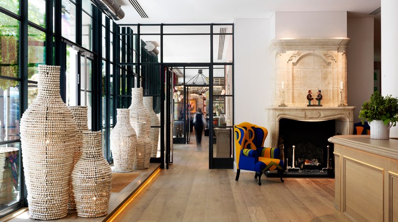A light and airy reception area with floor to ceiling windows, fronted by large, quirky, designed urns as art installations on either side of the glassed entrance doors, the corner of a reception desk on the right as well as two bright armchairs surrounding a fireplace.