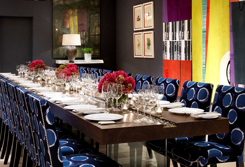 Rectangular wooden table set up for dining in a private event space, with comfortable dark blue chairs and lighter blue circles on, original artwork on the walls and bright flowers as centerpieces on the table.