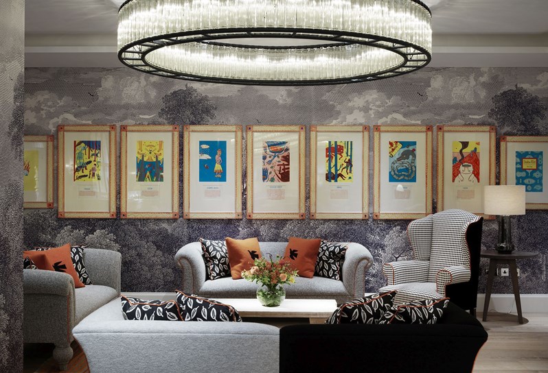Seating area of the Windmill private event room, with three comfortable sofas and an armchair around a coffee table, all in color schemes matching the dusky wallpaper, brightened up by an intricate circular chandelier in the ceiling and bold, colorful artworks on the walls.