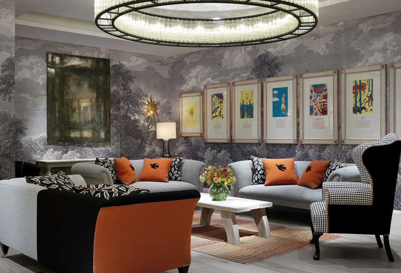 A side angle of the seating area in the Windmill private room, with three sofas and an armchair in grey, black and orange colors arranged around a low wooden coffee table and interesting art and a large mirror on the walls.