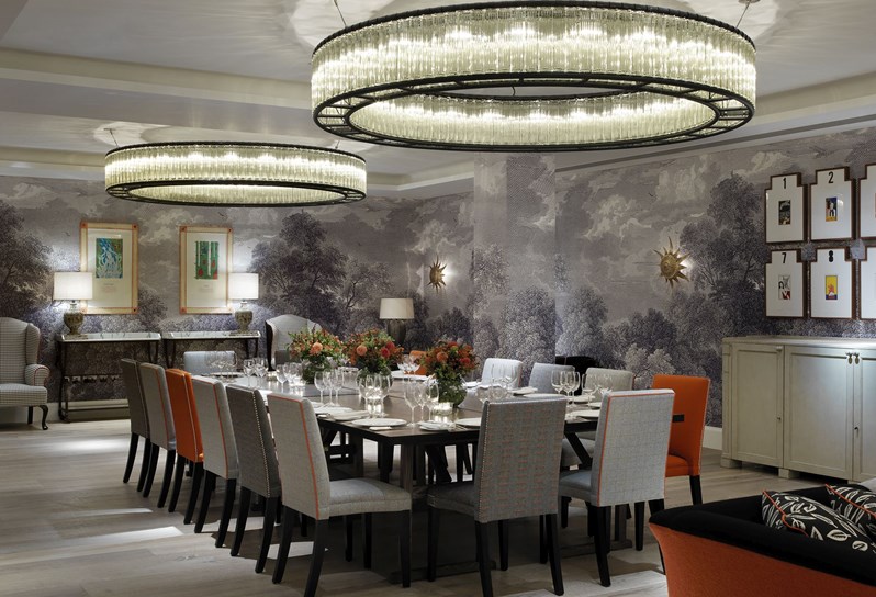 Large rectangular table set for private dining of 16 people with orange, light grey and dark grey comfortable chairs, matching flower arrangements as centerpieces, in the surrounds of the intricate light fixtures and bold artwork in the Windmill private event space.
