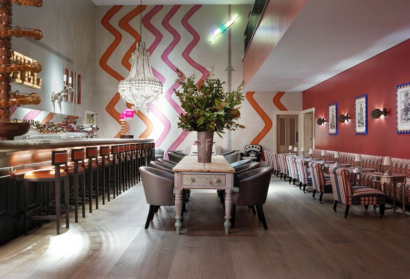 A view straight to a wall with Kit Kemp's signature Rick Rack wallpaper, with a fully stocked bar with seating on the left, low lounge furniture along the wall on the right and a statement wooden table with comfortable chairs around in the middle.