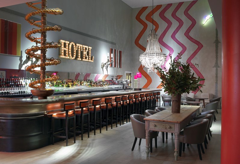 Angled view of a fully stocked bar with high chairs in front and two wooden tables with comfortable chairs on the side. There is a spiral art installation from the floor above filled with oranges ending in a fruit bowl on the bar, as well as colorful and playful artwork and wallpaper on the walls.