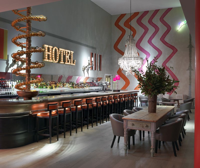 Angled view of a fully stocked bar with high chairs in front and two wooden tables with comfortable chairs on the side. There is a spiral art installation from the floor above filled with oranges ending in a fruit bowl on the bar, as well as colorful and playful artwork and wallpaper on the walls.