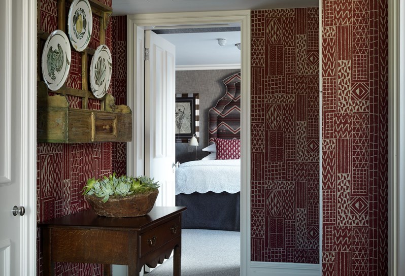 A shot of the hallway leading to a bedroom. There is a burgundy patterned wallpaper with a console table and decorative plates on the wall to the left. The bed is visible in the center of the photo.