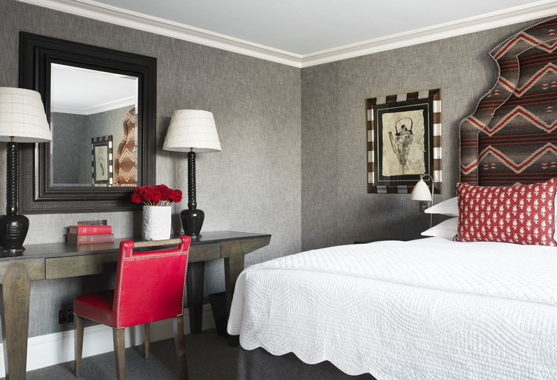 Side angle of a bedroom with grey walls and a bed with a dark patterned headboard and red cushions. There is a desk with a cerise chair, mirror hanging above and 2 lampshades on.