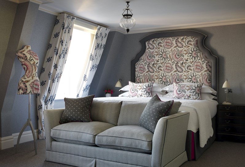 A grey bedroom with a sofa at the bottom of the bed and mannequin to the left, next to the window.