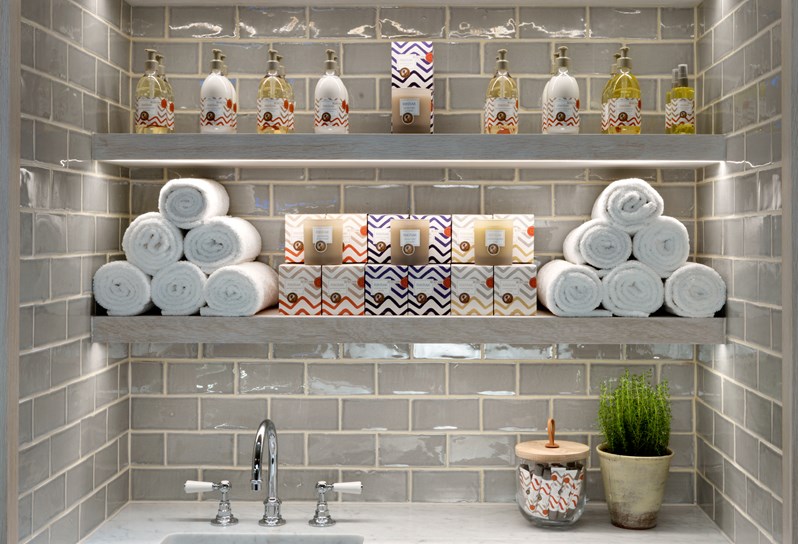 White mable sink with white shelved above. On the shelves are fresh white towels and Rik Rak products. On the right there is a small green plant.