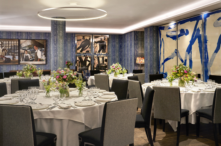 An image of multiple round dining table set up in the Indigo Room, there are white table cloths, floral bouquets in the centre and candles. each place around the table has a placemat, plate, cutlery, glassware and napkins ready for dining. private events room at The Soho Hotel.