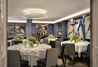 An image of multiple round dining table set up in the Indigo Room, there are white table cloths, floral bouquets in the centre and candles. each place around the table has a placemat, plate, cutlery, glassware and napkins ready for dining. private events room at The Soho Hotel.