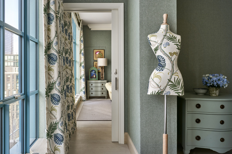 A Landscape image of a mannequin next to a corridor to the bedroom in a room in Warren Street Hotel. The scheme of the mannequin and the room is light blue with florals. 