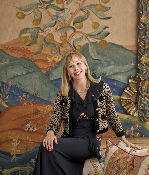A portrait image of Kit Kemp in front of a wall covered in Kit Kemp x Andrew Martin Meadow Hillside, in the Drawing Room at The Soho Hotel