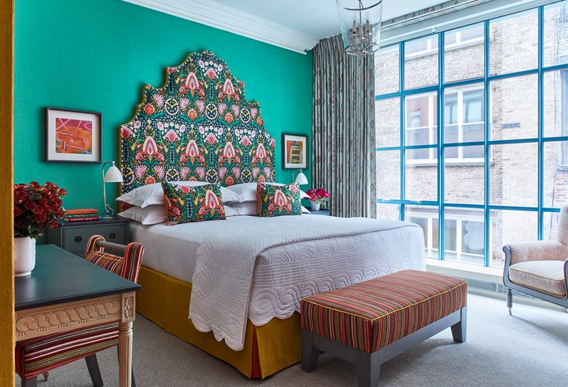 Red flowers on a desk with a striped chair. A bed with a multicoloured headboard, a striped ottoman and an orange and white armchair.