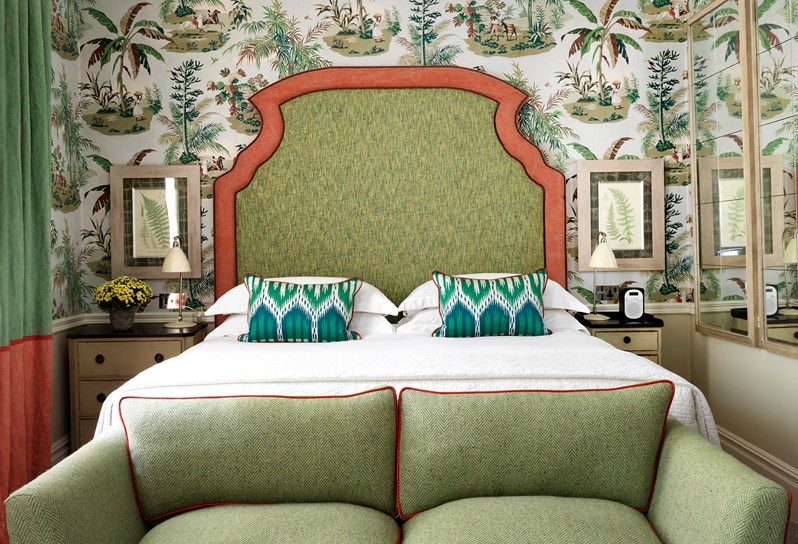 Front on bed shot in Room 111, at Covent Garden Hotel. There is a large green and orange headboard, curtains, sofa as well as a tropical themed wallpaper.
