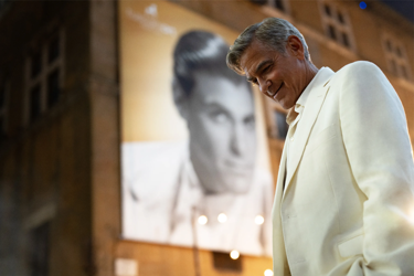 Still from the film Jay Kelly. A grey haired man dressed in a white suit looks down towards to floor smiling