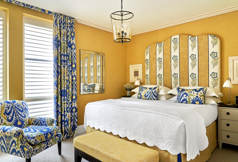 A yellow room facing a large bed with patterned headboard. A comfortable armchair sits below the window streaming light in to the left and there is an ottoman at the end of the bed.