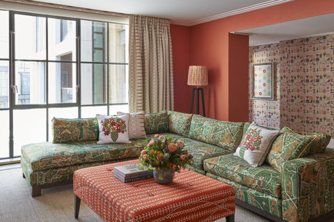 A living room space of the Deluxe Two Bedroom Suite Room 5 at Ham Yard Hotel.  A vibrant living room with red-orange walls, patterned green furniture, and eclectic décor floral arrangements, and framed textile art above the fireplace.