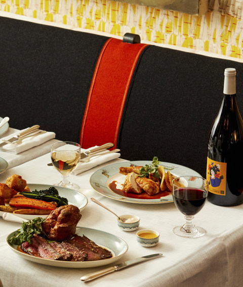An image to promote the Long Lunch at Ham Yard Bar & Restaurant at Ham Yard Hotel. The image shows a white-linen table set with an abundant spread of elegantly plated dishes, including roast beef with Yorkshire pudding, rolled pastry-wrapped meat, roast chicken, seasonal vegetables, and creamy mashed potatoes. Glasses of red and white wine are paired with the meal, with two bottles of wine displayed at either end of the table.