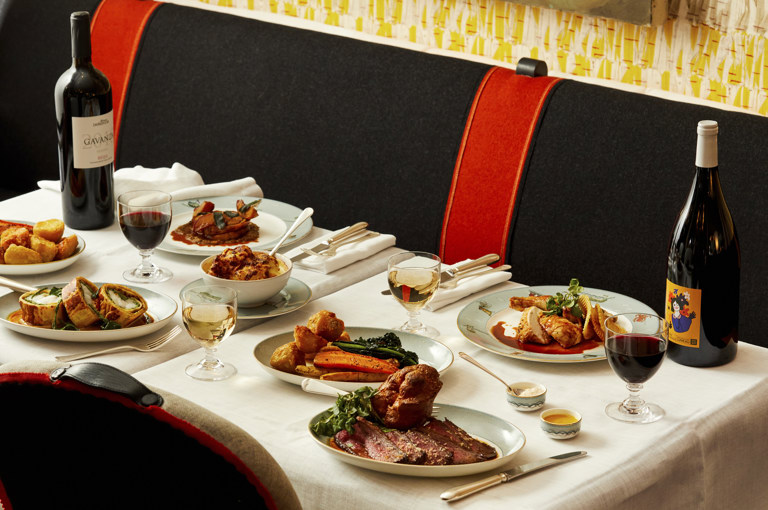 An image to promote the Long Lunch at Ham Yard Bar & Restaurant at Ham Yard Hotel. The image shows a white-linen table set with an abundant spread of elegantly plated dishes, including roast beef with Yorkshire pudding, rolled pastry-wrapped meat, roast chicken, seasonal vegetables, and creamy mashed potatoes. Glasses of red and white wine are paired with the meal, with two bottles of wine displayed at either end of the table.