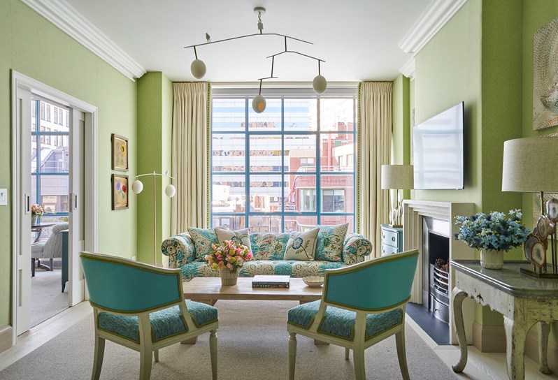 A green walled sitting room. Two blue and green Whitby chairs on one side of a coffee table, and a blue and green floral patterned sofa on the opposite side. Double doors open to the next room on the left and a fireplace and TV are on the right, with two tables and lamps either side.