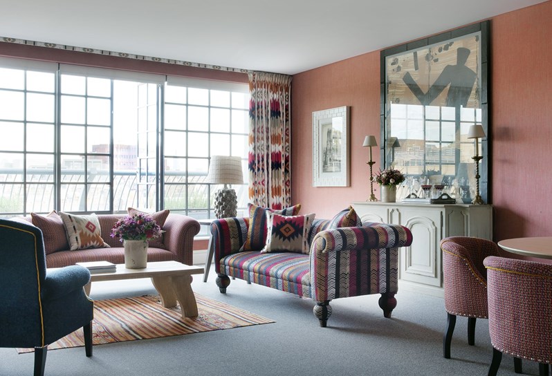 A living room in warm oranges, reds and pinks. There is a sitting area with two couches with patterned pillows and a chair, a coffee table in front of them sits on an orange striped carpet. In the background floor to ceiling windows lead to a terrace. To the right of the living space a large piece of art hangs above a mantel.