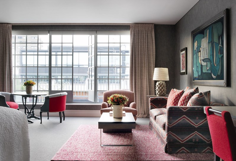 A view towards the windows showing the sitting area in a suite. There are pops of red in the neutral room. A brown couch with red details is on the right and in the foreground is a red chair, all sitting on a pink carpet. There is a sitting area towards the windows with a table and two red chairs. On the wall behind the couch is a blue abstract painting.