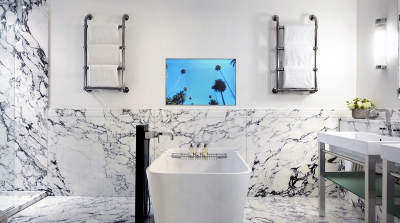A deep soaking tub in the middle of a bathroom with white Carrera marble. On the far wall is a television and two heated towel racks on either side. To the left is the entrance to the standing shower and to the right of the bathtub are two basins and flowers.